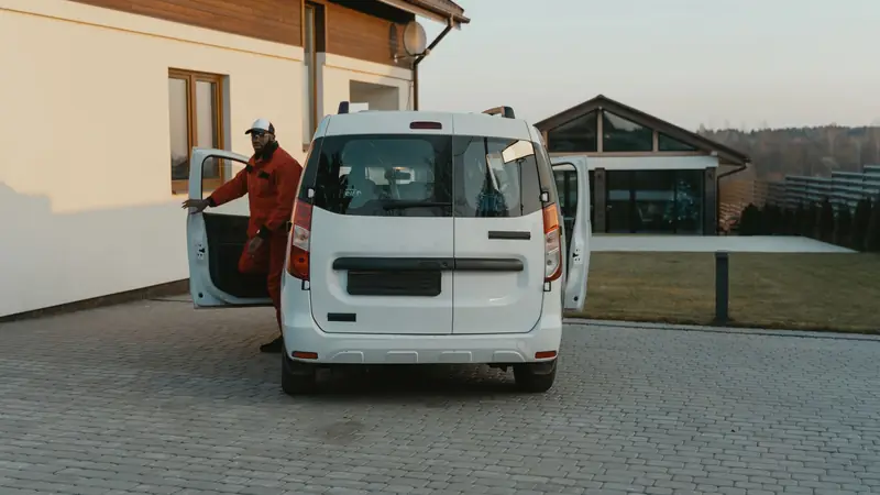 A worker stepping out of a white service van in a paved yard by a house